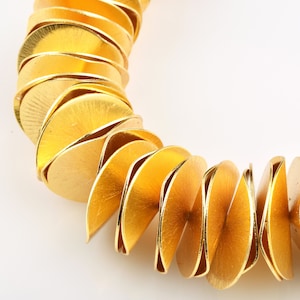 May include: A gold-colored necklace composed of overlapping, semi-circular metal pieces. The necklace has a brushed finish, and the design creates a layered, textured appearance. The jewelry is set against a white background, highlighting the warm tones of the metal.