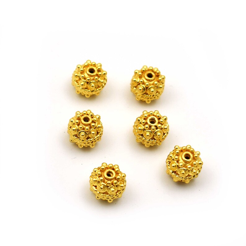 May include: Six gold-colored, spherical beads with a textured, dotted surface. Each bead has a central hole and is surrounded by smaller, raised dots. The beads are arranged on a white background.