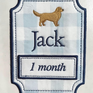 May include: Embroidered patch with a light blue and white gingham pattern. Features a golden retriever dog, the name "Jack", and the text "1 month" in navy blue. The patch has a navy blue border.