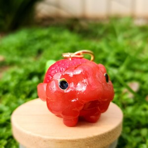 Puede incluir: Un peque&ntilde;o amuleto de arcilla polim&eacute;rica hecho a mano, de color rojo brillante, con forma de animal de dibujos animados con ojos grandes. El amuleto tiene un lazo dorado para sujetarlo a un llavero o collar.