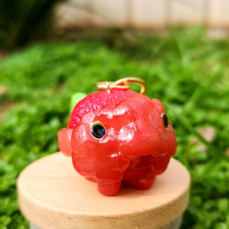 Puede incluir: Un peque&ntilde;o amuleto de arcilla polim&eacute;rica hecho a mano, de color rojo brillante, con forma de animal de dibujos animados con ojos grandes. El amuleto tiene un lazo dorado para sujetarlo a un llavero o collar.