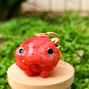 Puede incluir: Un llavero de pez rojo de resina con ojos negros. El llavero est&aacute; unido a un anillo de metal dorado.