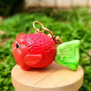 Puede incluir: Un amuleto de arcilla rojo y verde con forma de fresa con un lazo dorado para colgar.