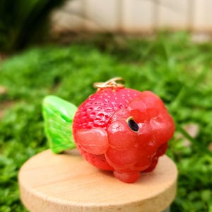 Puede incluir: Un amuleto de fresa de color rojo y verde con un anillo dorado. El amuleto est&aacute; hecho de arcilla polim&eacute;rica y tiene un aspecto realista.