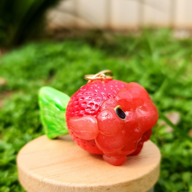 Puede incluir: Un amuleto de fresa de color rojo y verde con un anillo dorado. El amuleto est&aacute; hecho de arcilla polim&eacute;rica y tiene un aspecto realista.