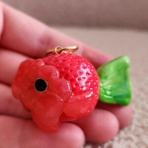 Puede incluir: Un amuleto en forma de fresa roja y verde con un lazo dorado. El amuleto est&aacute; hecho de arcilla polim&eacute;rica y tiene una textura detallada.