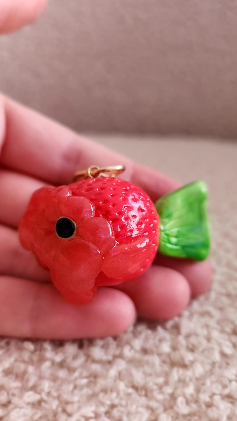 Puede incluir: Un amuleto en forma de fresa roja y verde con un lazo dorado. El amuleto est&aacute; hecho de arcilla polim&eacute;rica y tiene una textura detallada.
