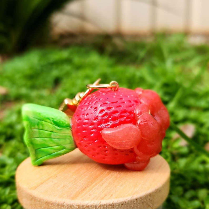 Puede incluir: Un amuleto de arcilla polim&eacute;rica roja y verde con forma de fresa con una cadena dorada unida. La fresa tiene una hoja verde y un cuerpo rojo con una superficie texturizada.