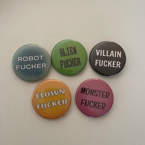 May include: Five round pinback buttons with the phrases "ROBOT FUCKER", "ALIEN FUCKER", "VILLAIN FUCKER", "CLOWN FUCKER", and "MONSTER FUCKER" printed in white on various colored backgrounds. The buttons are teal, green, black, orange, and pink.