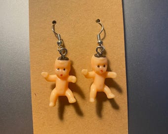 Op de afbeelding: Nieuwigheidsoorbellen met kleine, perzik kleurige plastic babyfiguren. De baby's hebben uitgestrekte armen en zijn opgehangen aan zilverkleurige oorhaakjes. De oorbellen worden gepresenteerd op een bruine kartonnen oorbellen displaykaart.