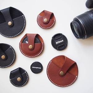 May include: A collection of handmade leather camera lens pouches in black and brown, with white stitching and brass snap closures. Includes Nikon and Olympus lens caps and a Nikon camera lens.