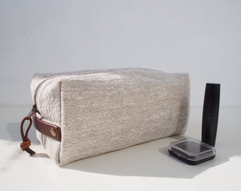 May include: A rectangular beige linen toiletry bag with a coarse woven texture, featuring a brown leather side handle secured with a brass rivet, a leather zipper pull, and a metal zipper. Next to it sits a black lipstick tube and a small compact.