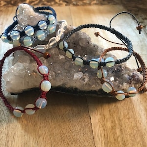 May include: Four bracelets with white opal beads and different colored cords. The cords are red, brown, dark blue, and light blue.