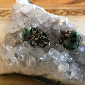 May include: Two rings with green gemstone settings on a bed of white crystals. The rings have an ornate, filigree design.