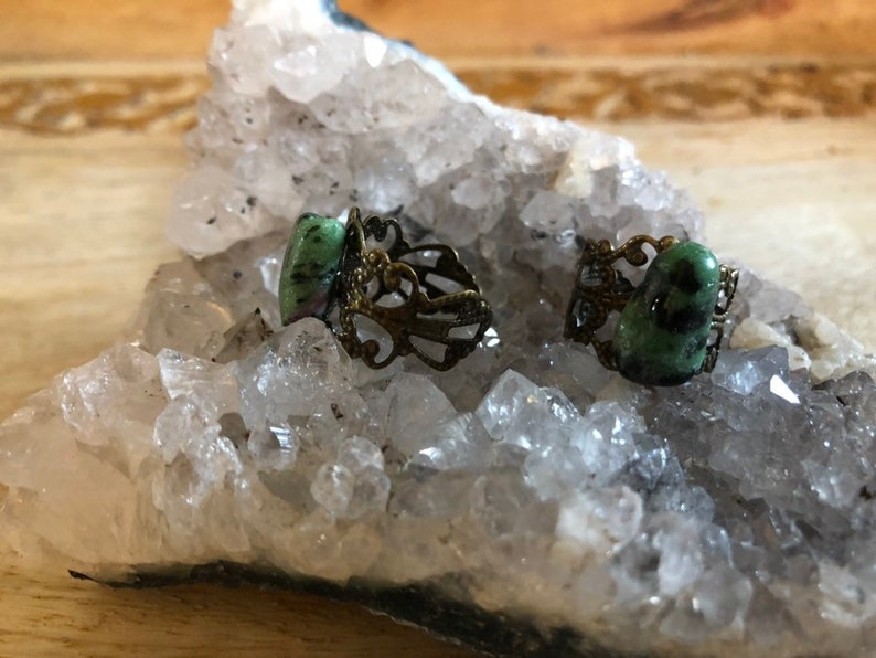 May include: Two rings with green gemstone settings on a bed of white crystals. The rings have an ornate, filigree design.