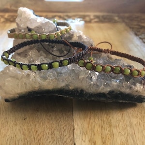 May include: Three macrame bracelets with green beads are displayed on a white crystal formation. The bracelets are in shades of brown and tan, with the green beads adding a pop of color. The bracelets are handmade.