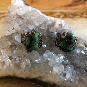 May include: Two green gemstone rings with ornate gold settings. The rings are displayed on a bed of white crystals.