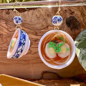 May include: A pair of unique earrings featuring miniature bowls of ramen. Each earring has a gold-colored hook, a blue and white floral bead, and a small bowl with ramen noodles, broth, and toppings. The bowls have a white base with blue floral patterns.
