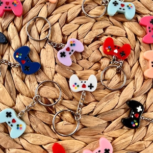 May include: A collection of colorful video game controller keychains. The keychains are in various colors, including pink, blue, red, and black. Each controller has a silver keyring and chain. The controllers have colorful buttons and directional pads.