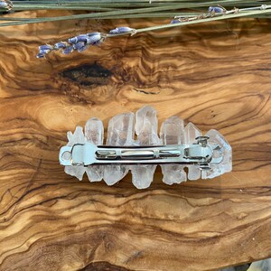 Op de afbeelding: Een haarspeld van helder kwartskristal met een zilveren metalen sluiting. De speld ligt op een houten oppervlak.