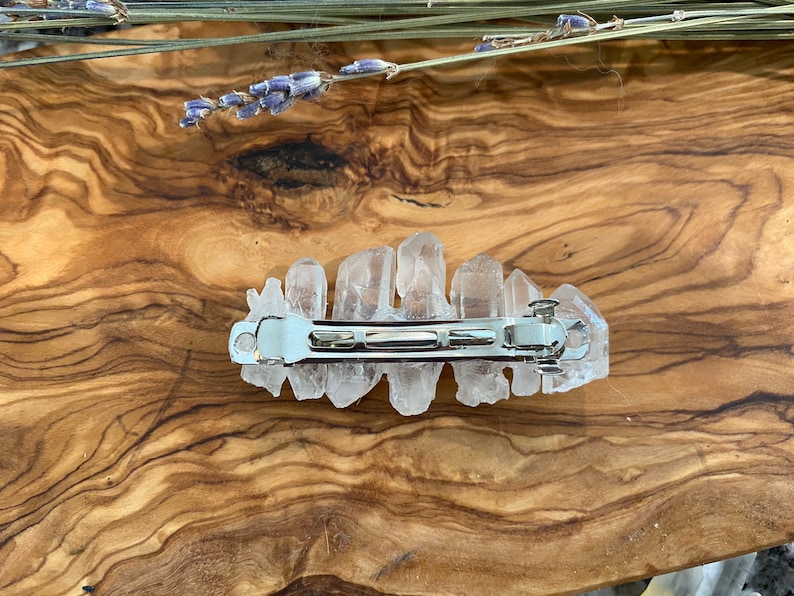 Op de afbeelding: Een haarspeld van helder kwartskristal met een zilveren metalen sluiting. De speld ligt op een houten oppervlak.