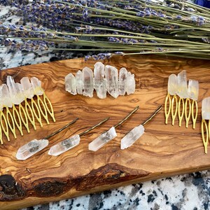 Op de afbeelding: Een set haar kammen en pinnen met heldere kwartskristallen. De kammen zijn goudkleurig en hebben een decoratief ontwerp. De pinnen zijn eenvoudig en recht. De kristallen zijn in verschillende maten en vormen.