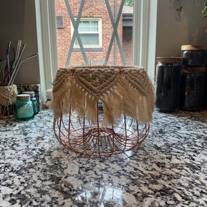 May include: A copper wire fruit basket with a beige macrame cover with fringe. The basket is sitting on a granite countertop.