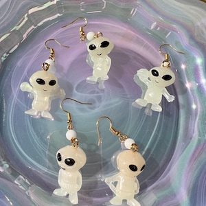 May include: Five pairs of dangle earrings featuring white alien figurines with black eyes. The earrings have gold hooks and white beads.