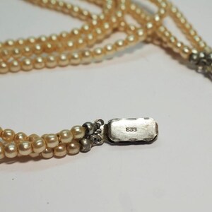 Puede incluir: Collar de perlas de varias hebras con cierre plateado. Las perlas son de color blanco crema. El cierre es rectangular y tiene el n&uacute;mero "835" estampado. El collar est&aacute; sobre una superficie blanca.