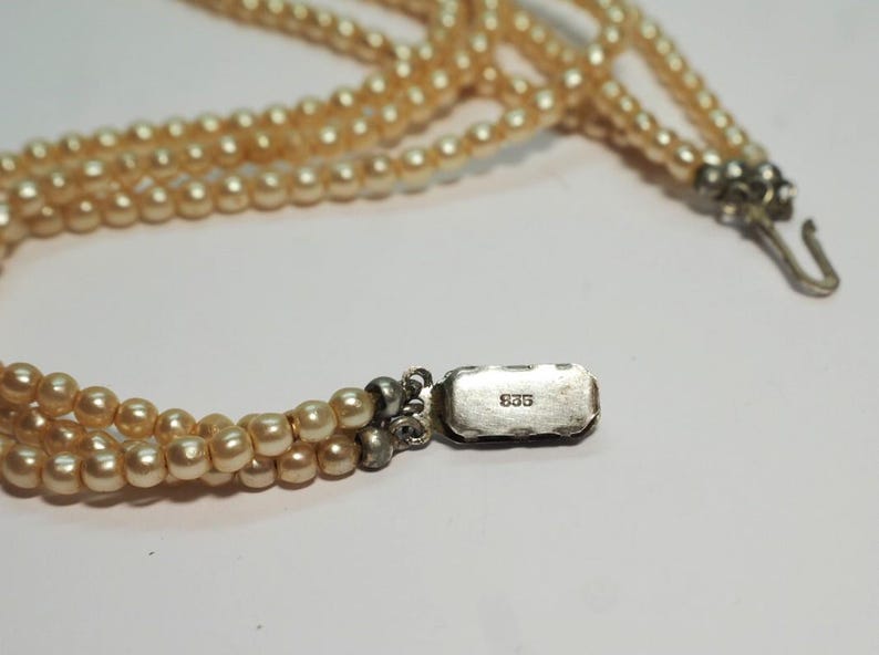 Puede incluir: Collar de perlas de varias hebras con cierre plateado. Las perlas son de color blanco crema. El cierre es rectangular y tiene el n&uacute;mero "835" estampado. El collar est&aacute; sobre una superficie blanca.