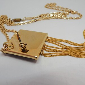 May include: A gold-toned necklace featuring a square pendant with a small key charm and cascading chain fringe. The necklace has a delicate chain and a clasp closure. The design is elegant and suitable for various occasions.
