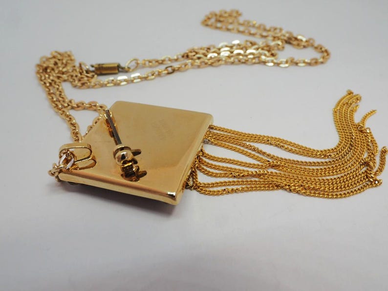 May include: A gold-toned necklace featuring a square pendant with a small key charm and cascading chain fringe. The necklace has a delicate chain and a clasp closure. The design is elegant and suitable for various occasions.
