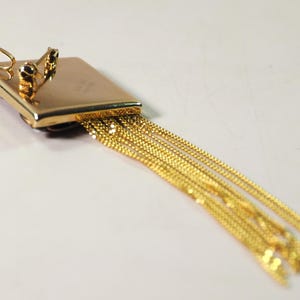 May include: A gold-toned pendant with a rectangular top and multiple dangling chains. The pendant has a small loop at the top and two decorative elements. The chains are made of small, linked segments.