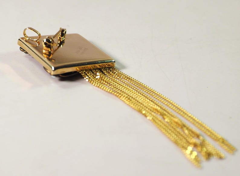 May include: A gold-toned pendant with a rectangular top and multiple dangling chains. The pendant has a small loop at the top and two decorative elements. The chains are made of small, linked segments.