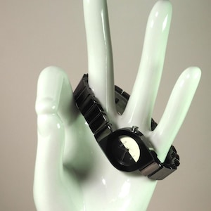 May include: A black wristwatch with a geometric face and a matching black band. The watch is displayed on a white, hand-shaped sculpture. The watch face features a white and black design.