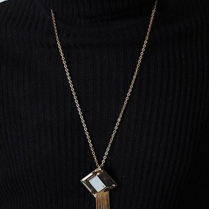 May include: A gold-toned necklace with a geometric pendant. The pendant is a square shape with a mirrored center and gold fringe. The necklace is worn over a black turtleneck.