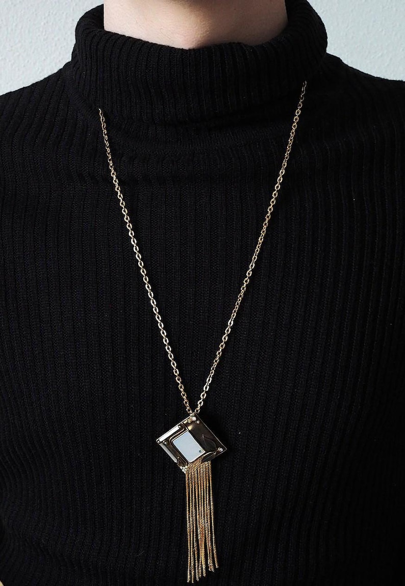 May include: A gold-toned necklace with a geometric pendant. The pendant is a square shape with a mirrored center and gold fringe. The necklace is worn over a black turtleneck.