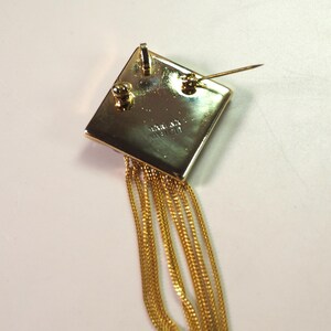 May include: A gold-toned brooch with a square top and dangling chains. The square has a pin on the back for attachment. The chains are thin and hang down from the bottom of the square. The brooch is likely vintage.