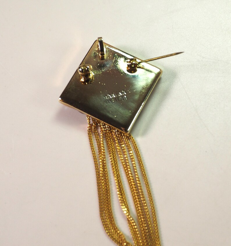 May include: A gold-toned brooch with a square top and dangling chains. The square has a pin on the back for attachment. The chains are thin and hang down from the bottom of the square. The brooch is likely vintage.