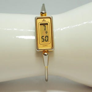 May include: A rectangular digital watch with a gold-toned face and a silver-toned metal bangle band. The watch face displays the time 7:50 in black digital numerals, a small star icon, a crown logo at the top, and the word QUARZ at the bottom.