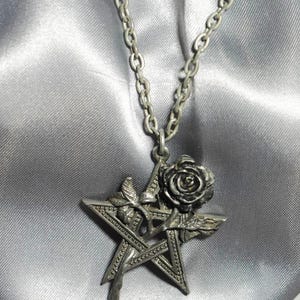 May include: Silver-toned pendant necklace featuring a pentagram design with a rose and leaf accents. The pendant hangs from a delicate chain. The necklace is displayed against a silver satin background.