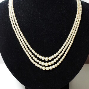 Puede incluir: Un collar de perlas de tres hilos exhibido sobre un busto de terciopelo negro. Las perlas son de color blanco roto, con tama&ntilde;os variables en cada hilo. El collar est&aacute; drapeado en una curva suave, mostrando su elegante dise&ntilde;o.