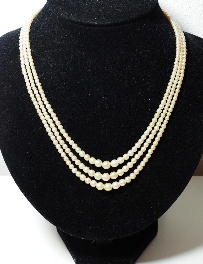 Puede incluir: Un collar de perlas de tres hilos exhibido sobre un busto de terciopelo negro. Las perlas son de color blanco roto, con tama&ntilde;os variables en cada hilo. El collar est&aacute; drapeado en una curva suave, mostrando su elegante dise&ntilde;o.