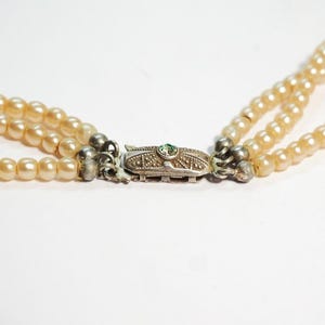 Puede incluir: Collar de perlas de tres hilos con cierre plateado. Las perlas son de color crema c&aacute;lido. El cierre es rectangular con una peque&ntilde;a piedra verde en el centro. El collar est&aacute; sobre un fondo blanco.