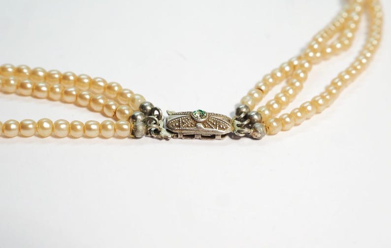 Puede incluir: Collar de perlas de tres hilos con cierre plateado. Las perlas son de color crema c&aacute;lido. El cierre es rectangular con una peque&ntilde;a piedra verde en el centro. El collar est&aacute; sobre un fondo blanco.