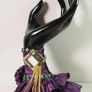 May include: A black hand-shaped jewelry display holding a purple dress with a gold and diamond-accented brooch. The dress has a pleated texture and a black belt. Gold chains hang from the brooch.