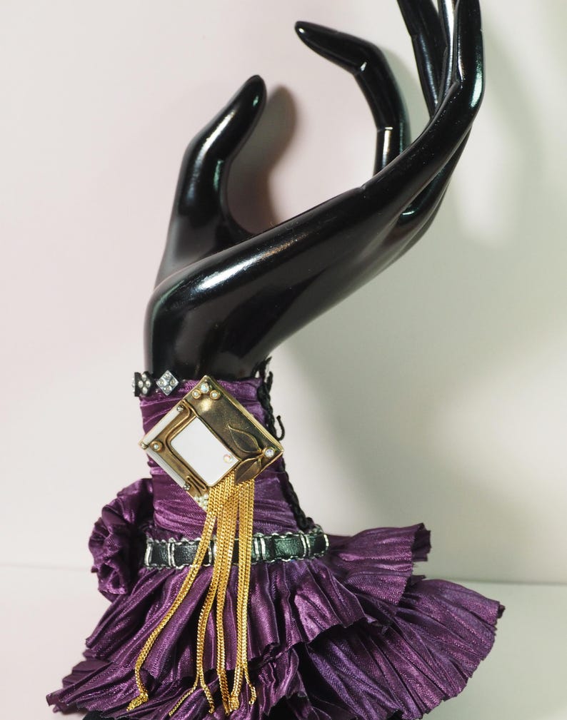 May include: A black hand-shaped jewelry display holding a purple dress with a gold and diamond-accented brooch. The dress has a pleated texture and a black belt. Gold chains hang from the brooch.