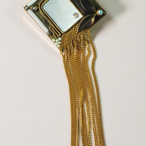 May include: An ornate, vintage-style brooch with a rectangular mirror at its center. The brooch features a gold-toned frame with leaf and pearl accents, and dangling gold chains. The design is Art Deco inspired.