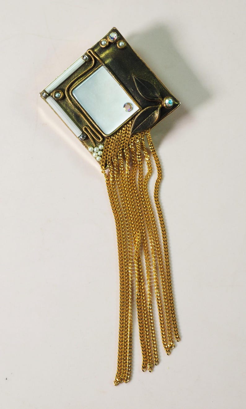 May include: An ornate, vintage-style brooch with a rectangular mirror at its center. The brooch features a gold-toned frame with leaf and pearl accents, and dangling gold chains. The design is Art Deco inspired.