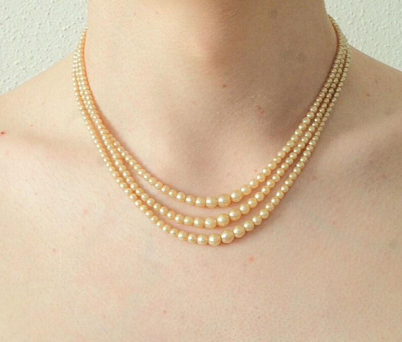 Puede incluir: Un collar de perlas de tres hilos con un suave color crema. El collar est&aacute; compuesto por perlas graduadas, con las perlas m&aacute;s grandes en el centro y las m&aacute;s peque&ntilde;as hacia el cierre. Una joya cl&aacute;sica.